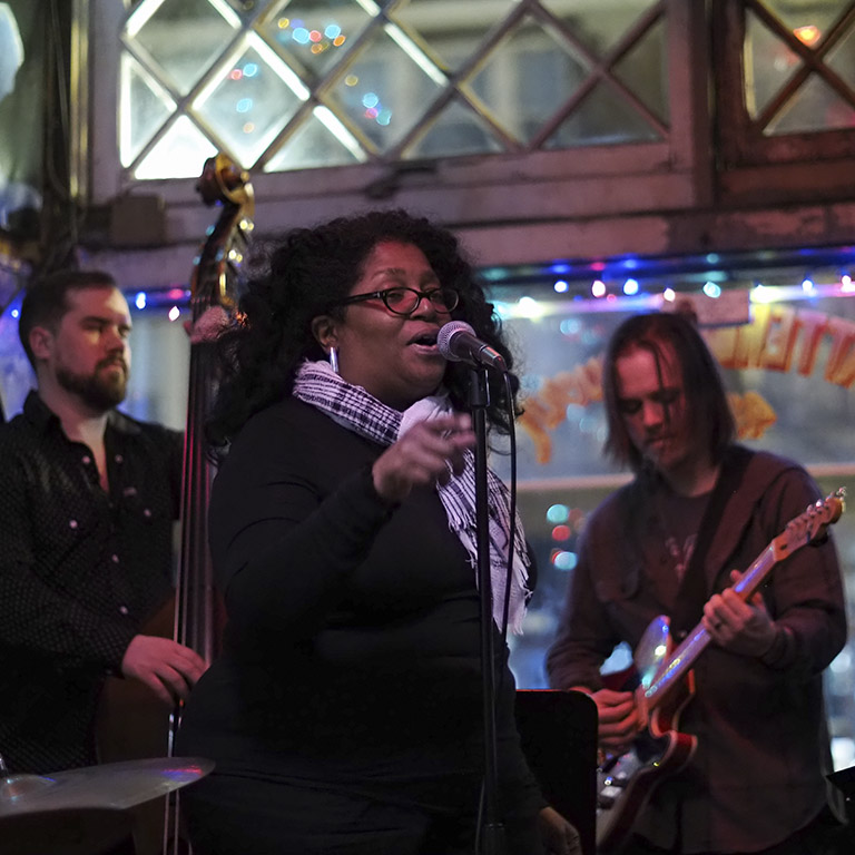 Woman singing with an upright bass player and guitar player in the background at the Chatterbox