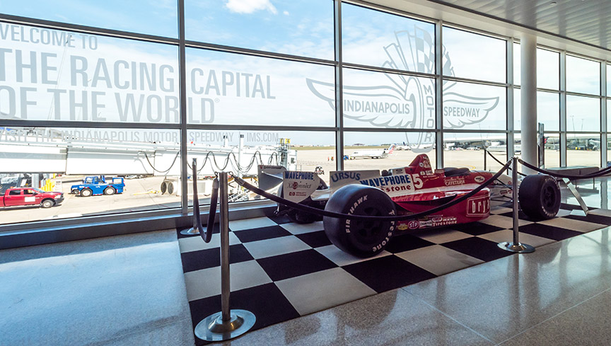 An Indy Car on display in front of a window with text proclaiming Racing Capital of the World with the Indianapolis Motor Speedway logo