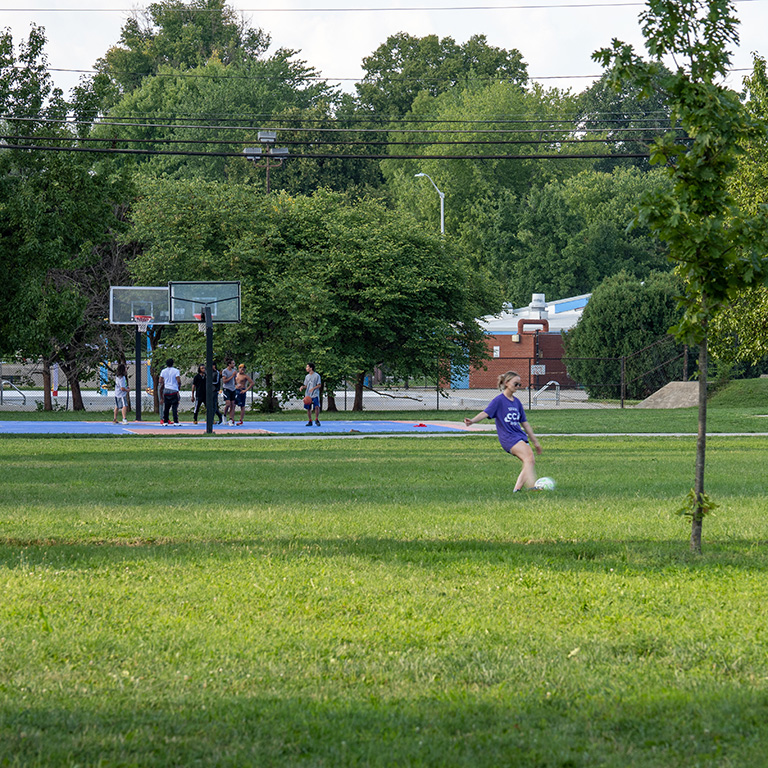 Young woman kicks a soccer ball with young men playing basketball in the background.